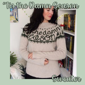 Op de afbeelding: Beige trui met de tekst "'Tis the Damn Season" en "Sweater" in een scriptlettertype. De trui heeft een ronde hals en lange mouwen. De tekst is donkergroen. De trui is een neutrale kleur.