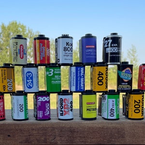 Pu&ograve; includere: Una pila di 15 diverse cassette di pellicola, disposte a forma di piramide. Le cassette sono di marchi diversi e hanno diverse sensibilit&agrave; ISO, tra cui 200, 400 e 800. Le cassette sono appoggiate su una superficie di legno.