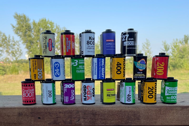 Pu&ograve; includere: Una pila di 15 diverse cassette di pellicola, disposte a forma di piramide. Le cassette sono di marchi diversi e hanno diverse sensibilit&agrave; ISO, tra cui 200, 400 e 800. Le cassette sono appoggiate su una superficie di legno.