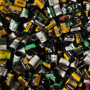 Pu&ograve; includere: Un'immagine ravvicinata di una variet&agrave; di cassette di pellicola in vari colori e dimensioni. Le cassette sono per lo pi&ugrave; gialle e nere con testo e numeri bianchi. Alcune delle cassette hanno il nome del marchio "Kodak" stampato su di esse.