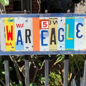 May include: A wooden sign with the word "WAR EAGLE" spelled out using license plates from different states. The sign is painted blue and has a distressed look.