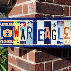 May include: A blue wooden sign with the word "WAREAGLE" spelled out using various license plates. The license plates have different state names and logos, including "Auburn Tigers", "Mississippi", and "Texas".