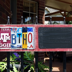 May include: A red wooden sign with a chalkboard on the right side. The left side of the sign is decorated with license plates spelling out "TAMBTHO" with the letters "A" and "M" in maroon, "B" in blue, "T" in yellow, and "H" and "O" in red. The license plates also include the text "Texas A&M University" and "Aggie E. Longhorn S.A."