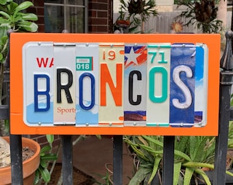 May include: A wooden sign with an orange background and the word "BRONCOS" spelled out using licence plates. The plates are from Washington state and have the numbers 19, 71, and 018.