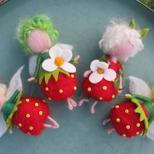 Op de afbeelding: Vier grillige vilten aardbeienfee-ornamenten. Elke fee heeft een roze gezicht, witte vleugels en een aardbeienlichaam met gele zaden. De feeën hebben groen, wit en geel haar en dragen witte bloemenhoeden.