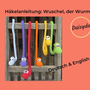 May include: A crochet pattern for a colorful, long, worm-shaped toy. The pattern is available in both German and English. The image shows seven crocheted worms hanging on a wooden fence. The worms are in the colors blue, purple, red, orange, yellow, and green.