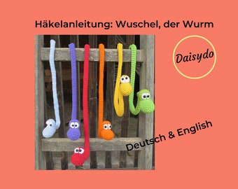 May include: A crochet pattern for a colorful, long, worm-shaped toy. The pattern is available in both German and English. The image shows seven crocheted worms hanging on a wooden fence. The worms are in the colors blue, purple, red, orange, yellow, and green.