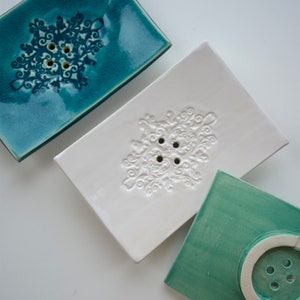 Könnte beinhalten: Drei Keramik-Seifenschalen mit dekorativen Designs. Die erste Schale ist blaugrün mit einem weißen Schneeflockenmuster. Die zweite Schale ist weiß mit einem weißen Schneeflockenmuster. Die dritte Schale ist hellgrün mit einem weißen Kreisdesign.