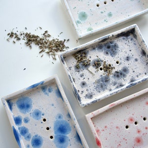 Könnte beinhalten: Vier Keramik-Seifenschalen mit verschiedenen gesprenkelten Mustern. Die Schalen sind weiß mit blauen, grünen, grauen und roten Sprenkeln. Jede Schale hat drei Abflusslöcher.