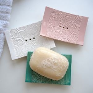 Op de afbeelding: Drie keramische zeepbakjes in wit, roze en teal, elk met een paisley patroon. Een stuk zeep met bubbels ligt op het teal bakje. De rechthoekige bakjes hebben twee afvoergaten.