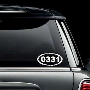 May include: A white oval sticker with the text "0331 Machine Gunner" on a black car window.