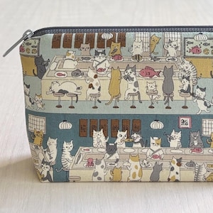 May include: A rectangular, zippered pouch with a whimsical print of cats in a sushi restaurant. The fabric features a light blue background with various cats, sushi, and restaurant details. The zipper is silver.