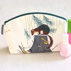 May include: A cream-colored cosmetic bag with a forest scene featuring a brown animal on a tree stump. The bag has a green zipper and a gray pull tab. Two bottles of nail polish are next to the bag.