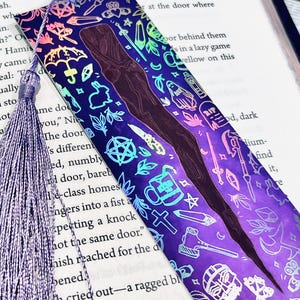 May include: A holographic bookmark with a purple background and a rainbow iridescent finish. The bookmark features a variety of illustrations, including a witch, a cauldron, a knife, a cross, a skull, and a crescent moon. The bookmark is designed to be used with a book.