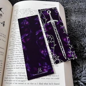 K&ouml;nnte beinhalten: Zwei Lesezeichen mit einem violetten Blumenmuster und einem wei&szlig;en Schwertdesign. Das Lesezeichen mit dem Schwertdesign enth&auml;lt den Text "Sophie Elaina Designs" in wei&szlig;en Buchstaben.