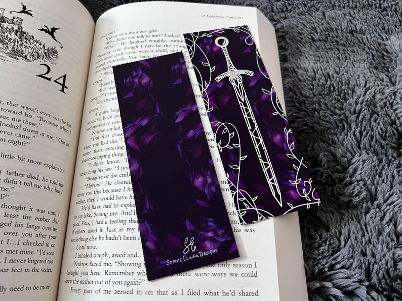 K&ouml;nnte beinhalten: Zwei Lesezeichen mit einem violetten Blumenmuster und einem wei&szlig;en Schwertdesign. Das Lesezeichen mit dem Schwertdesign enth&auml;lt den Text "Sophie Elaina Designs" in wei&szlig;en Buchstaben.