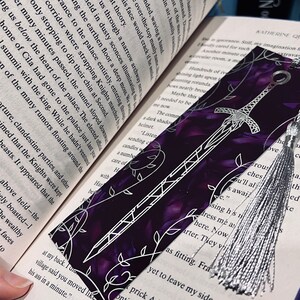 K&ouml;nnte beinhalten: Ein violettes und silbernes Lesezeichen mit einem silbernen Quasten. Das Lesezeichen zeigt ein silbernes Schwertdesign mit einem floralen Muster auf einem violetten Hintergrund.