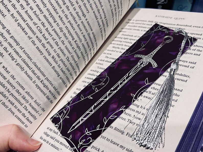 K&ouml;nnte beinhalten: Ein violettes und silbernes Lesezeichen mit einem silbernen Quasten. Das Lesezeichen zeigt ein silbernes Schwertdesign mit einem floralen Muster auf einem violetten Hintergrund.