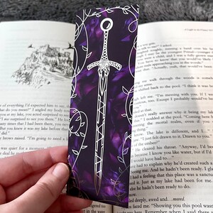 K&ouml;nnte beinhalten: Ein violettes und silbernes Lesezeichen mit einem Schwertmuster. Das Schwert ist in Silber umrandet und hat einen violetten Hintergrund. Das Lesezeichen ist von einem silbernen Rankenmuster umgeben.