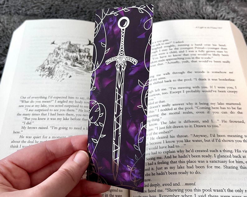 K&ouml;nnte beinhalten: Ein violettes und silbernes Lesezeichen mit einem Schwertmuster. Das Schwert ist in Silber umrandet und hat einen violetten Hintergrund. Das Lesezeichen ist von einem silbernen Rankenmuster umgeben.