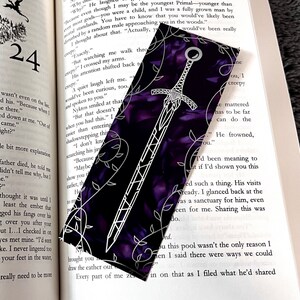 K&ouml;nnte beinhalten: Ein violettes Lesezeichen mit einem wei&szlig;en Schwertdesign und floralen Akzenten. Das Schwert hat einen schwarzen Griff und eine silberne Klinge.