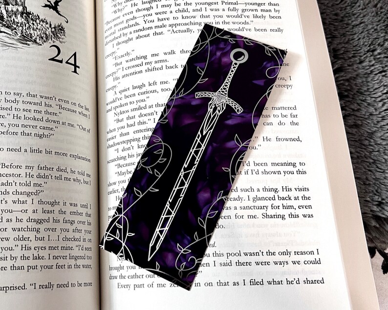 K&ouml;nnte beinhalten: Ein violettes Lesezeichen mit einem wei&szlig;en Schwertdesign und floralen Akzenten. Das Schwert hat einen schwarzen Griff und eine silberne Klinge.