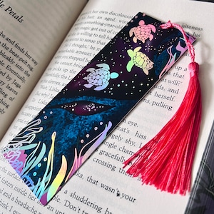 May include: A decorative bookmark with a sea turtle design and a pink tassel. The bookmark has a dark blue and purple background with white stars and sea plants. The turtles are in shades of pink, yellow, and green.