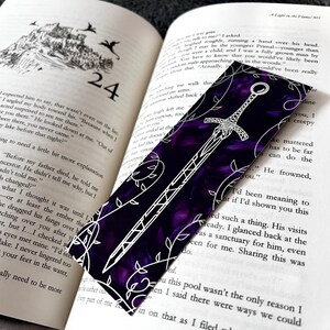 K&ouml;nnte beinhalten: Ein violettes und wei&szlig;es Lesezeichen mit einem Schwertmuster. Das Lesezeichen ist aus einem strukturierten Material gefertigt und hat ein Blumenmuster.
