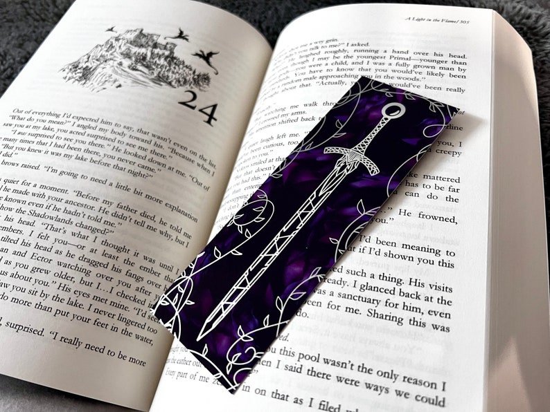 K&ouml;nnte beinhalten: Ein violettes und wei&szlig;es Lesezeichen mit einem Schwertmuster. Das Lesezeichen ist aus einem strukturierten Material gefertigt und hat ein Blumenmuster.