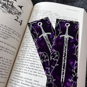 K&ouml;nnte beinhalten: Zwei violette Lesezeichen mit einem wei&szlig;en Schwertmuster und einem Rankenmuster. Die Schwerter sind wei&szlig; umrandet und haben ein detailliertes Design. Der Hintergrund ist dunkelviolett mit einem dezenten Blumenmuster.