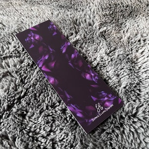 K&ouml;nnte beinhalten: Ein violettes Blumen-Lesezeichen mit dem Text "S.E. Designs" in wei&szlig;er Schrift.