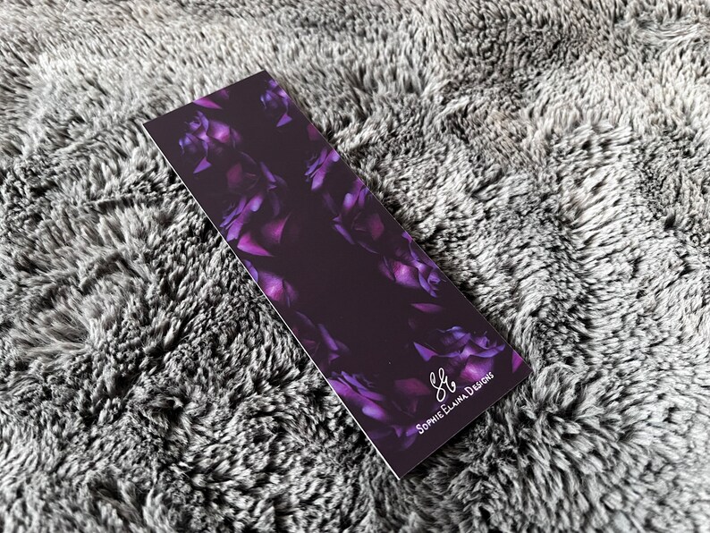K&ouml;nnte beinhalten: Ein violettes Blumen-Lesezeichen mit dem Text "S.E. Designs" in wei&szlig;er Schrift.