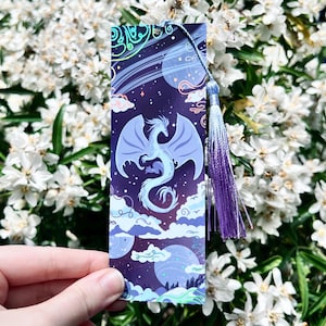 May include: A fantasy-themed bookmark featuring a blue dragon with outstretched wings against a starry night sky background. The bookmark has a purple tassel and is held in front of a backdrop of white flowers.