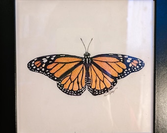 May include: A framed artwork featuring a monarch butterfly. The butterfly has orange and black wings with white spots, set against a white background. The artwork is enclosed in a black frame, suitable for wall display.