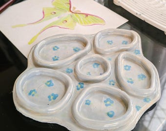 May include: A ceramic artist's palette with multiple wells, decorated with blue floral patterns. A watercolor painting of a green and yellow butterfly and a white paint brush are nearby. A white, textured paint brush cleaner is also present.