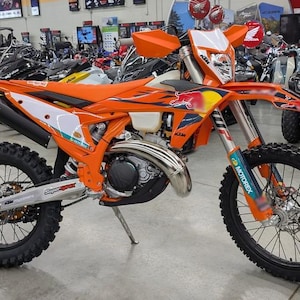 May include: An orange and white KTM dirt bike with black tires. The bike has a Red Bull logo and Motorex stickers. Other vehicles are visible in the background.