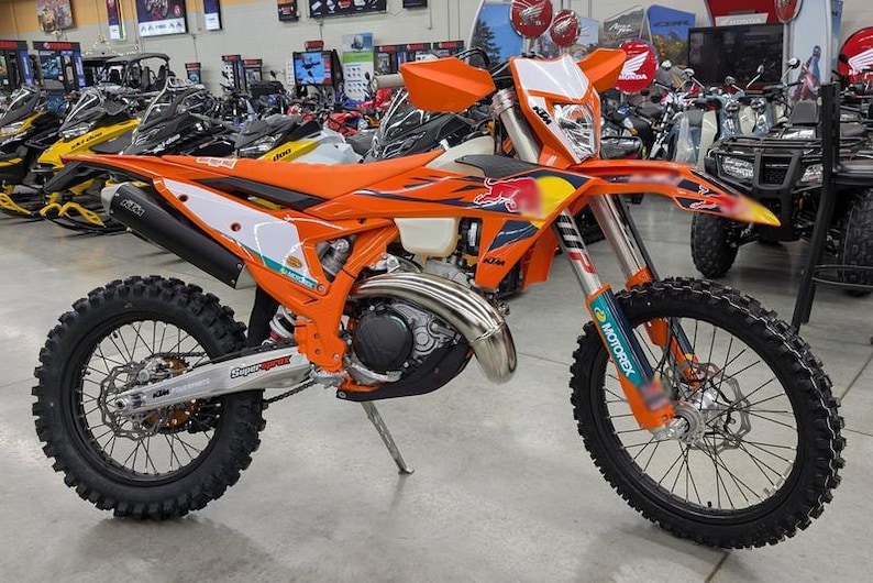 May include: An orange and white KTM dirt bike with black tires. The bike has a Red Bull logo and Motorex stickers. Other vehicles are visible in the background.