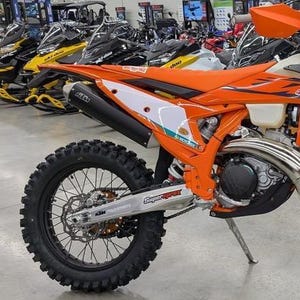 Könnte beinhalten: Ein orange-weißes Dirtbike mit schwarzen Reifen und einem schwarzen Auspuff. Das Motorrad hat eine silberne Schwinge mit dem Text "SuperSprox" und "KTM PowerParts". Im Hintergrund sind weitere Schneemobile und Motorräder zu sehen.