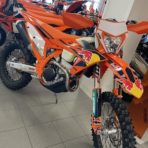 May include: An orange and white KTM dirt bike with Red Bull branding. The motorcycle has a black seat, black tires, and a white headlight. Other orange KTM motorcycles are visible in the background.