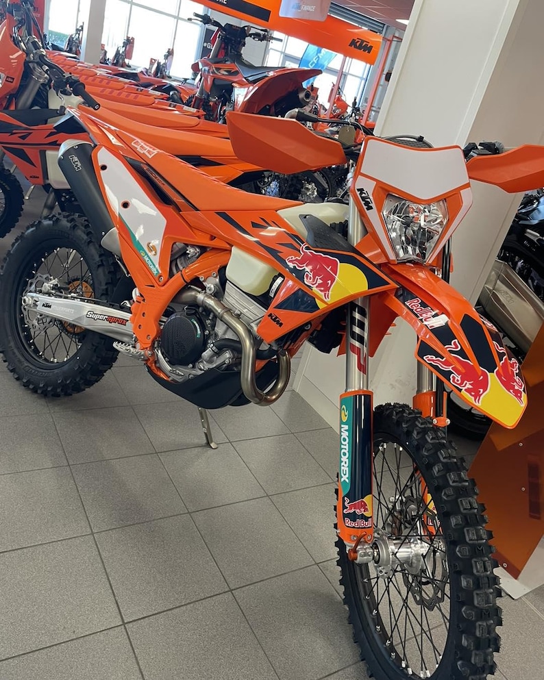 May include: An orange and white KTM dirt bike with Red Bull branding. The motorcycle has a black seat, black tires, and a white headlight. Other orange KTM motorcycles are visible in the background.