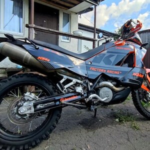 May include: A black and orange KTM motorcycle featuring Factory Racing branding, knobby tires, a high-mounted exhaust, and visible Brembo and Renthal components. The bike has a sleek frame with orange accents and a black seat, designed for off-road use.