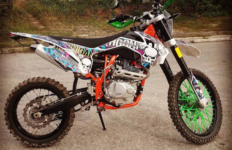 May include: A black and orange dirt bike with green spoke covers on the front wheel. The bike has a white and black graphic design with the word "Holligan" on the side.