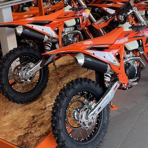 May include: Several orange and white dirt bikes are displayed, showcasing their sleek design. The bikes feature black tires, silver accents, and black exhaust pipes. The KTM logo is visible on the bikes, indicating the brand.