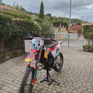 May include: A red, white, and orange motocross motorcycle with the number 28 on the front and side panels. The motorcycle has black tires and is on a black stand. The background includes a brick driveway and green foliage.