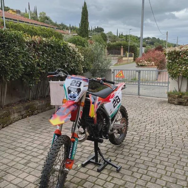 May include: A red, white, and orange motocross motorcycle with the number 28 on the front and side panels. The motorcycle has black tires and is on a black stand. The background includes a brick driveway and green foliage.