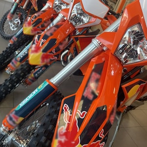 May include: Close-up of several orange and white dirt bikes. The bikes have silver front forks, black tires, and the Red Bull logo. The image is taken indoors, with the bikes lined up in a row.