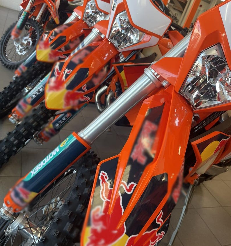 May include: Close-up of several orange and white dirt bikes. The bikes have silver front forks, black tires, and the Red Bull logo. The image is taken indoors, with the bikes lined up in a row.