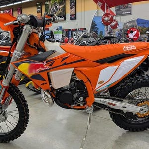 May include: An orange and black dirt bike with a Red Bull logo on the side. The motorcycle has a black tire, a white fuel tank, and a silver exhaust pipe. The bike is in a showroom.