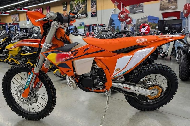 May include: An orange and black dirt bike with a Red Bull logo on the side. The motorcycle has a black tire, a white fuel tank, and a silver exhaust pipe. The bike is in a showroom.