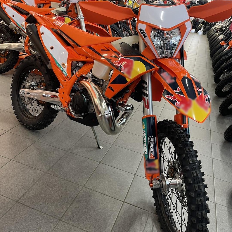 May include: An orange and white KTM dirt bike with black tires and a silver exhaust pipe. The bike has a Red Bull logo on the front and side panels. The motorcycle is in a showroom.